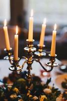 Shot of antique candlestick with five burning candles stand on table against light blurred background with floral arrangement. Lit candles in dining room photo