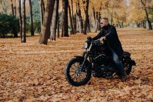 Horizontal outdoor view of active male motorcyclist rides bike, wears trendy sunglasses and black jacket, poses in big autumn park, enjoys awesome travel, has motorcycle trip, rest at nature. photo