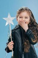 Overemotive positive small child covers mouth with one palm, wears fashionable black leather jacket and crown, holds magic wand in hand, isolated over blue background. Female kid poses indoor photo