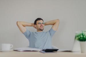 Carefree male enterpreneur ready with financial report, being glad to realize that he recieves high net profit, sits at working desk with calculator, laptop computer, documentation. Accounting concept photo