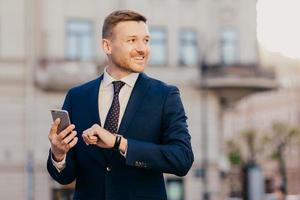 Horizontal shot of successful unshaven male CEO looks for interesting multimedia files on smart phone, downloads or updates new programs, stands against blurred city background, looks happily aside photo