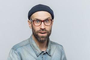 Headshot of good looking unshaven male with blue eyes, looks with suprised expression into camera, feels excited and shocked with latest news, poses against grey background. Surprisment and people photo