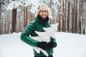 Attractive bearded male with appealing appearance, wears green anorak and hat, holds artificial fir tree, spends weekends on frosty weather in winter forest. Winter, weather and lifestyle concept photo