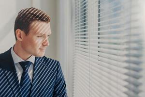 Indoor shot of pensive male economist dressed in formal outfit, looks pensively at window with jalousie conemplates about work during dinner break feels tired after working. Business, people, thoughts photo