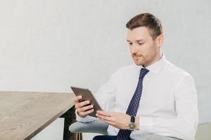 Horizontal shot of handsome young successful male fiinancier uses modern tablet computer, wears white shirt with tie, has break after work, sits at wooden table in his cabinet, reads information photo
