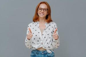 Positive redhaired woman with gentle smiles shows thumbs up, demonstrates like or approval gesture, says well done, likes someting, wears polka dot shirt and jeans, isolated on grey studio wall photo