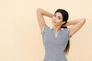 Portrait of pretty female with dark hair, makes pony tail, dressed in casual striped t shirt, looks seriously at camera, poses against studio wall with blank space for your advetisement or promotion photo