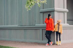 Horizontal view of fashionable European female in red loose sweater, ripped jeans, holds hands together with her younger sister, pose outside together, have walk in open air, have friendly relations photo