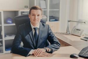 Half length portrait of handsome prosperous male manager sits at cabinet, looks directly into camera, has meeting with colleagues, discuss new business strategy. People and occupation concept photo