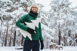 Cheerful male model dressed in winter clothes, holds white artificial fir tree, stands in winter forest, has pleasant smile, enjoys beautiful landscapes. People, holidays, recreation concept photo