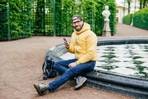 Smiling happy man with bristle wearing casual clothes and big eyewear sitting crossed legs near fountain in park holding cell phone messaging with his girlfriend having delightful expression photo