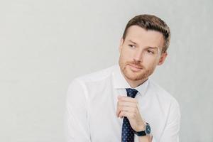 concepto de personas y confianza. atractivo joven empleado sin afeitar mira cuidadosamente a un lado, contempla algo importante, usa camisa blanca y corbata, aislado sobre fondo blanco foto