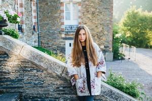Outdoor portrait of attractive woman with long luxurious hair, wears fashionable mantel, stands on ancient stairs of builduing, poses seriously at camera. People, lifestyle, tourism concept.