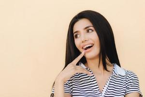 retrato de una joven feliz con cabello largo y oscuro, tiene una amplia sonrisa, indica en su boca, muestra dientes con frenos, se ve positivamente a un lado, aislada sobre el fondo del estudio. foto