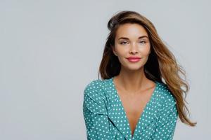 una foto de estudio de una joven positiva tiene el pelo largo y ondulado, maquillaje, usa una blusa de lunares turquesa, mira directamente a la cámara, modelos contra fondo blanco, copia espacio para su anuncio