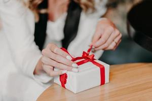 Photo of unrecognizable Caucasian woman ties red ribbon on small white gift box, prepares surprise for somebody on Chirstmas, New Year or birthday, blurred background. Close up, selective focus