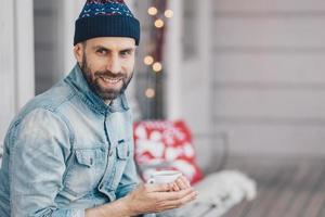 Portrait of handsome young smiling male enjoys spare time, holds mug of coffee, being deep in thoughts, has coffee break during shining warm day, focused on something. Pleased bearded stylish man photo