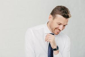 Positive unshaven man looks happily down, dressed in elegant clothes, stands against white background, isolated over white background with free space for advertising content. Happy male boss photo