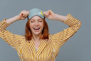 Overjoyed red haired Caucasian woman laughs positively, wears blindfold and yellow striped pajama, expresses good emotions, isolated on grey background. Good rest, bedtime and sleeping concept photo