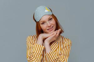 Portrait of good looking woman gets enough amount of sleep, enjoys pleasant relaxation, tilts head and keeps hands under chin, wears blindfold and striped pajamas, isolated over grey background photo