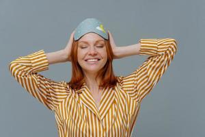 concepto de siesta durante el día. una mujer complacida con cabello pelirrojo usa un cómodo pijama a rayas y una máscara para dormir, usa un accesorio útil para dormir, sonríe ampliamente, muestra dientes blancos, aislado en la pared gris del estudio foto