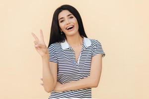 Waist up portrait of happy brunette young female shows peace sign, smiles happily at camera, wears braces on teeth, has friendly look, models indoor. People, body language and happiness concept photo