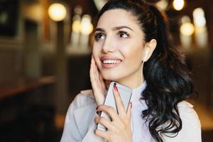 Fashion lifestyle concept. Portrait of cute woman with pony tail having charming smile listening to music with earphones using mobile phone while resting in cafe. Caucasian brunette female with mobile photo