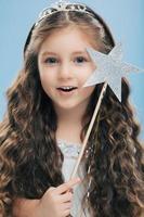Close up shot of adorable blue eyed small European female princess has long wavy hair, wears crown, holds star magic wand, isolated over blue studio background. Vertical image. Childhood concept photo
