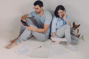 Young woman and man sit back to each other, do accounts together, make necessary calculations, surrounded with paper documents, sit on floor in empty room with dog. People and finances concept photo