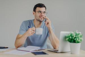 Pleasant looking male accountant in round glasses sips hot beverage, has mobile conversation, manages finances, works with laptop computer, writes notes in diary. Man answers call from bank. photo