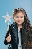 una niña pequeña satisfecha tiene el pelo largo y rizado, vestida con una chaqueta de cuero negro, lleva una corona, sostiene una varita mágica, aislada sobre un fondo azul. niño positivo de cabello oscuro anticipa un milagro foto