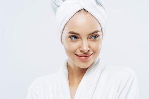 Day spa, beauty and freshness concept. Pensive European woman with healthy skin concentrated aside thoughtfully, feels refreshed after bath procedures, dressed in dressing gown, towel on head photo