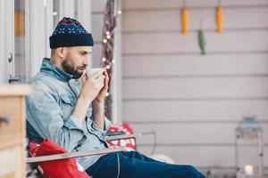 Photo of handsome male remembers something in life while drinks tea or coffee, has thoughtfull look down, recreats at terrace, wears fashionable hat and jean jacket. Leisure and people concept