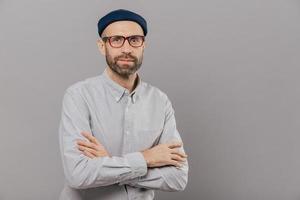 Photo of serious male keeps arms folded over chest, dressed in white formal shirt, fashionable hat, poses for making photography over grey wall with free space for your advertisement or promotion
