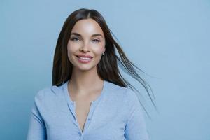 Positive lovely feminine lady happy to have first date with boyfriend, smiles broadly, has appealing appearance, glad to hear something pleasant, models indoor alone, wears blue casual jumper photo