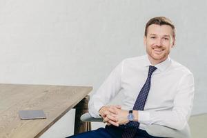 Horizontal shot of prosperous male entrepreneur in formal clothes, holds hands together, has positive expression, sits at wooden table with touch pad, waits for business partners to have meeting photo