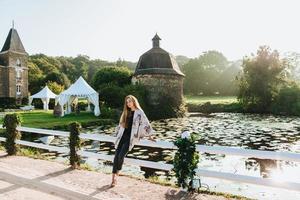 Thoughtful sad female in white cape stands near white short hence and small pond or lake, has walk on ancient historical territory, admires beautiful landscapes. People, lifestyle, excursion concept photo