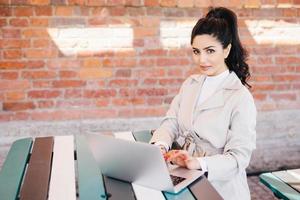 Portrait of cute female model having dark wavy pony tail wearing white elegant coat sitting at desk typing at computer translating article, looking at the camera with serious look posing on brick wall photo