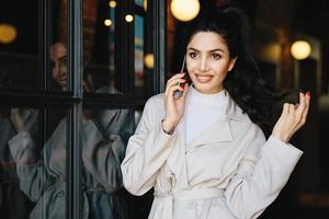 personas, estilo de vida, tecnologías modernas y concepto de moda. retrato de mujer de moda con cabello oscuro, ojos brillantes con pestañas largas y labios carnosos con chaqueta blanca usando un teléfono inteligente al aire libre foto