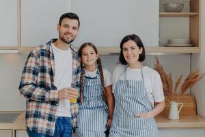 Happy family have great time together, pose in modern kitchen at home. Glad man in checkered shirt holds glass of juice, small child with pigtails, pretty housewife in apron. Girl with parents photo