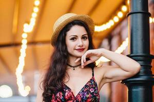 Hermosa mujer joven y elegante con apariencia atractiva posando en la terraza, vestida con sombrero de paja y vestido de verano, con la mano en la barbilla, con una mirada segura, mirando directamente a la cámara. foto