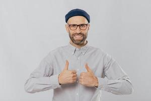 I like this idea Glad unshaven man raises thumbs up, smiles broadly, satisfied with great choice, has dark stubble, dressed in formal shirt, isolated over white background, approves cool idea photo