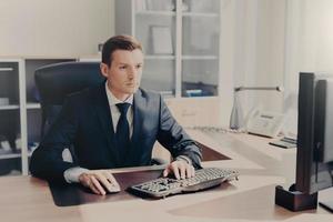 Serious concentrated businessman sits in front of computer at work place, keyboards information, wears suit, prepares business project for next year, poses in spacious cabinet. People and finances photo