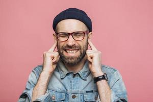 Annoyed bearded male plugs ears as hears unpleasant sound, keeps eyes shut, being dissatisfied with something, isolated over pink background. Unshaven middle aged male ignores hearing someone photo