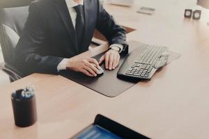 Cropped image of successful male business owner sits at work place in formal suit, uses computer for work, prepares financial report. Male executive works in office alone. People and finances photo