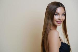 Portrait of pleased delighted woman with long dark hair, smiles gently, looks with attentive gaze at camera, poses against beige background, blank copy space aside. Party preparation, femininity photo