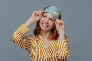 Dreamy redhaired European woman looks gladfully aside, smiles broadly, wears casual domestic clothes and sleepmask, isolated over grey background, imagines something pleasant. Good morning concept photo