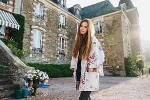 Portrait of atrrative woman wears white loose cape, stands outdoor against ancient builduing, has excursion in ancient city, has serious dreamful expression, looks directly in camera. Having nice day photo