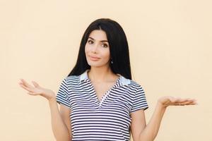 Shot of hesitant lovely female shrugs shoulders with puzzlement, doesnt know answer on difficult question, poses against light pink background. Clueless brunette woman has doubtful expression photo