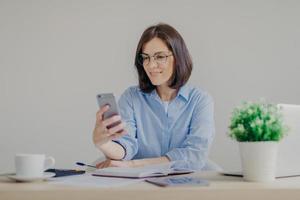 una exitosa mujer independiente morena recibe buenas noticias en un teléfono inteligente mientras trabaja en una computadora portátil, lee una publicación en el sitio web, toma notas en el diario, bebe café fuerte, hace algunos cálculos foto